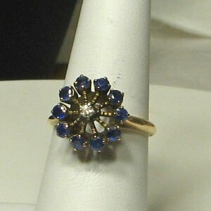 Pode incluir: Um anel de ouro com um grupo de safiras azuis e um &uacute;nico diamante no centro, formando uma flor.