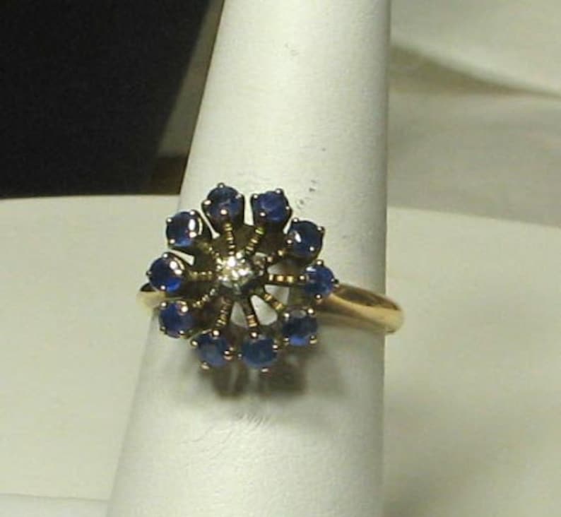 Pode incluir: Um anel de ouro com um grupo de safiras azuis e um &uacute;nico diamante no centro, formando uma flor.