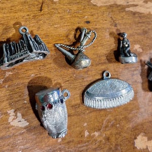 May include: A collection of small, silver-toned charms on a wooden surface. The charms include a mountain range with text, a teapot, a monkey, a windmill, a cup with red and blue accents, and a hat-shaped charm.