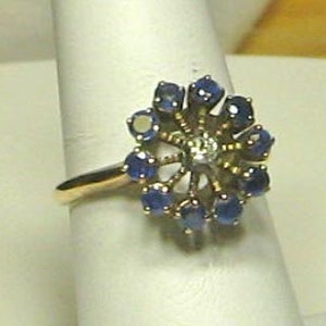 Pode incluir: Um anel de ouro com um grupo de safiras azuis e um &uacute;nico diamante no centro, formando um design floral.