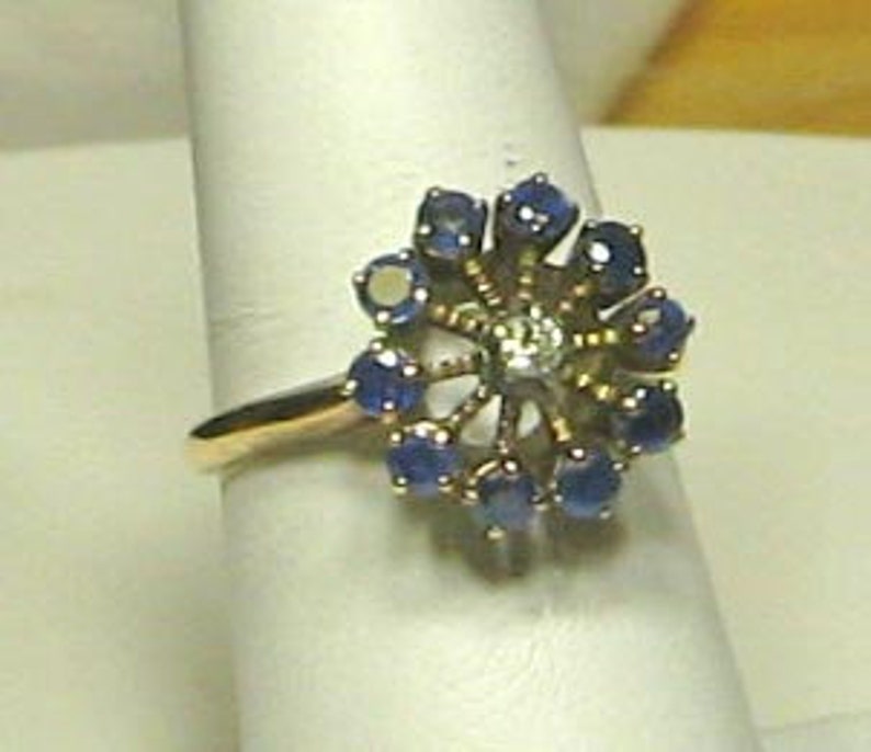 Pode incluir: Um anel de ouro com um grupo de safiras azuis e um &uacute;nico diamante no centro, formando um design floral.