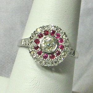 May include: A silver-toned ring featuring a central round-cut white stone surrounded by a halo of small, vibrant red rubies. An outer border of tiny, clear white stones encircles the rubies, with additional small stones set along the ring band.