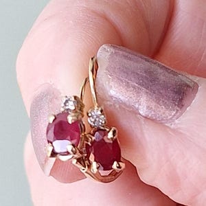 Puede incluir: Un par de pendientes dorados con cierre de palanca. Cada pendiente presenta una piedra preciosa roja oscura de forma ovalada y un pequeño acento de piedra preciosa transparente. Los pendientes están sostenidos por una mano con manicura rosa claro.