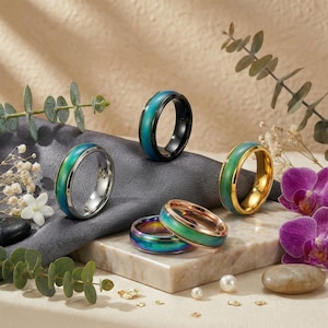 Può includere: Una collezione di anelli colorati con fasce iridescenti. Gli anelli presentano una gamma di colori, tra cui verde acqua, verde e oro, con fasce argentate, nere e dorate. Gli anelli sono esposti su uno sfondo neutro con accenti floreali.