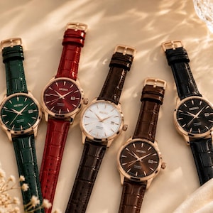 Op de afbeelding: Vijf polshorloges met leren bandjes in verschillende kleuren: groen, rood, bruin, wit en zwart. Elk horloge heeft een goudkleurige kast en een bijpassende gesp. De wijzerplaten tonen de merknaam Seiko.