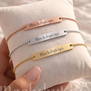 May include: Three personalized bar bracelets in rose gold, silver, and gold, each with the engraved text "MocksbyKiya". The bracelets are displayed on a white pillow, showcasing their delicate chain design and rectangular bar pendants.