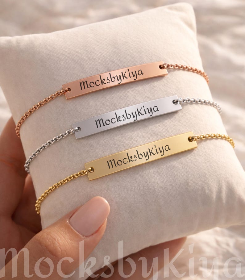 May include: Three personalized bar bracelets in rose gold, silver, and gold, each with the engraved text "MocksbyKiya". The bracelets are displayed on a white pillow, showcasing their delicate chain design and rectangular bar pendants.