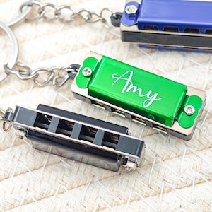 May include: Three miniature harmonicas in blue, green, and black, each attached to a keychain. The green harmonica has the name "Amy" in white script. The harmonicas are on a textured, off-white surface.