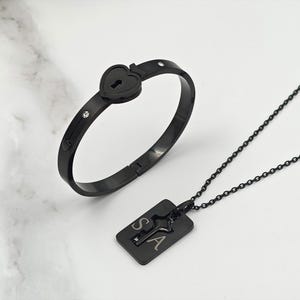 May include: Black matching jewelry set featuring a heart-shaped lock bracelet with three small crystals and a rectangular pendant necklace with the letters "S" and "A". The necklace has a chain.