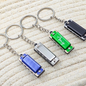 May include: Four miniature harmonica keyrings in black, green, silver, and blue. The green harmonica has the name "Amy" in white script. Each keyring has a silver chain and a silver key ring.