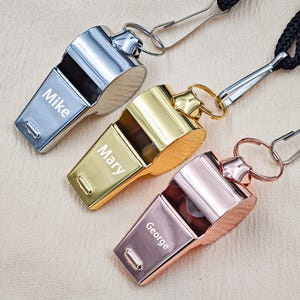 May include: Three personalized metal whistles in silver, gold, and rose gold. Each whistle has a name engraved on it: "Mike", "Mary", and "George". The whistles are attached to black lanyards with metal clips.