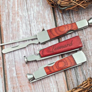 Puede incluir: Tres herramientas para pipas de tabaco con mangos de acero inoxidable y madera. Una herramienta está grabada con el nombre "Leonard" en cursiva. Las herramientas están dispuestas sobre una superficie de madera marrón claro.