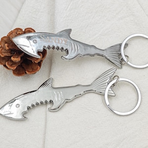 May include: Two silver shark-shaped bottle openers with key rings. One opener has the name "SANDY" engraved on it. The openers are lying on a white surface with a pine cone.