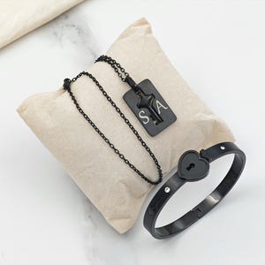Puede incluir: Un conjunto de joyas negro con un collar con un colgante rectangular y una pulsera. El colgante tiene un diseño de llave y las letras "SA". La pulsera tiene un candado en forma de corazón y pequeños cristales.