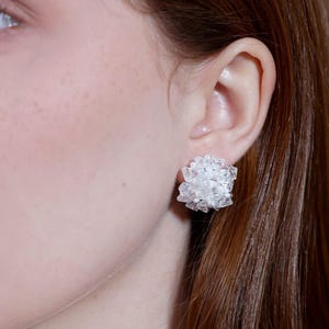 May include: Close-up of a clear, cluster-style stud earring. The earring is made of multiple small, irregularly shaped pieces, creating a textured, sparkling appearance. The earring is worn on the ear of a person with fair skin and reddish-brown hair.