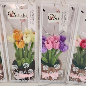 Può includere: Quattro bouquet di tulipani fatti a mano in sacchetti di plastica trasparente. Ogni bouquet presenta tulipani colorati nelle tonalità del rosa, arancione, giallo, viola e bianco, con steli e foglie verdi. I bouquet sono in vasi decorativi con fiocchi rosa.