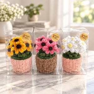 May include: Three clear plastic boxes each containing a crocheted flower pot. The left pot is pink with yellow sunflowers, the center is brown with pink flowers, and the right is pink with white daisies. Each box has a handmade with love sticker.