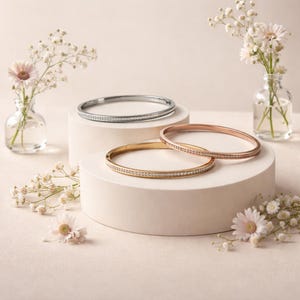 May include: Three circular bangle bracelets in silver, gold, and rose gold tones. Each bracelet features a thin band encrusted with a row of small, clear, sparkling stones along the outer surface. The bangles are arranged on two tiered platforms.