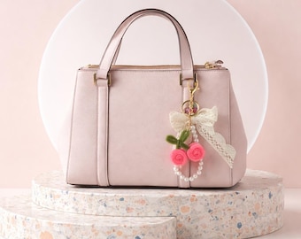May include: A light pink handbag with a gold zipper and handles. A decorative charm with pink roses, green leaves, and a white bow hangs from the handle. The bag is displayed on a multi-level, speckled, stone-like pedestal.