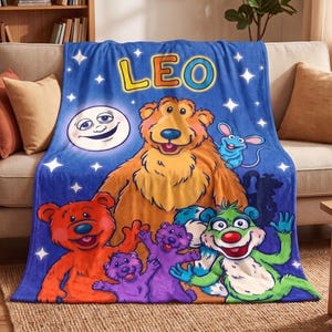 May include: A blue fleece blanket featuring the name LEO in yellow and orange letters, a smiling moon, and a group of colorful cartoon characters including a large brown bear, a red bear, purple creatures, and a green creature with stars.