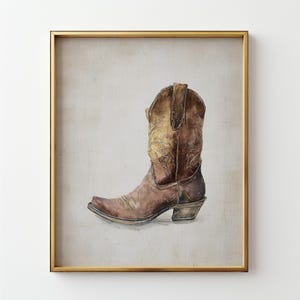 May include: A framed watercolor painting of a brown cowboy boot. The boot has intricate floral designs on the shaft and a pointed toe. The artwork is set against a neutral background and framed in a gold-colored frame.