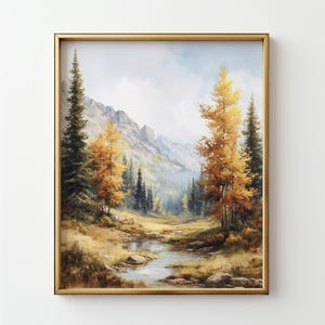 May include: A framed landscape painting depicts a serene mountain scene. The artwork features a stream flowing through a valley, surrounded by trees with golden and green foliage. The sky is a soft blue, and the painting is encased in a gold frame.
