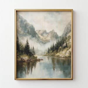 May include: A framed landscape painting depicts a serene mountain lake scene. The artwork features a gold frame, with a misty mountain range in the background, and evergreen trees lining the water's edge. The water reflects the sky and mountains.