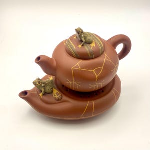 Puede incluir: Una tetera de cerámica marrón con una base a juego, con figuras de rana en la tapa y la base. La tetera tiene un asa y un pico curvos. La tapa y el cuerpo están decorados con líneas doradas.