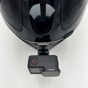 May include: A black motorcycle helmet with a camera mount attached. The camera is black with a red button and the brand name "NEEWER" is visible. The helmet is glossy and reflects the surrounding environment.