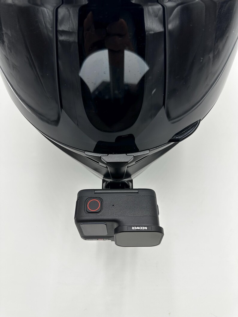 May include: A black motorcycle helmet with a camera mount attached. The camera is black with a red button and the brand name "NEEWER" is visible. The helmet is glossy and reflects the surrounding environment.