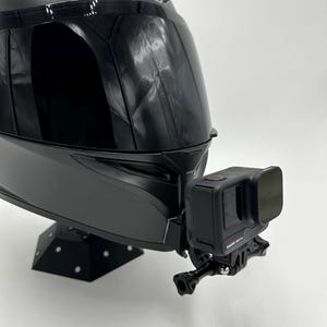 May include: A black motorcycle helmet with a mounted black action camera. The helmet has a glossy finish and the camera is attached to the front. The helmet is on a black stand.