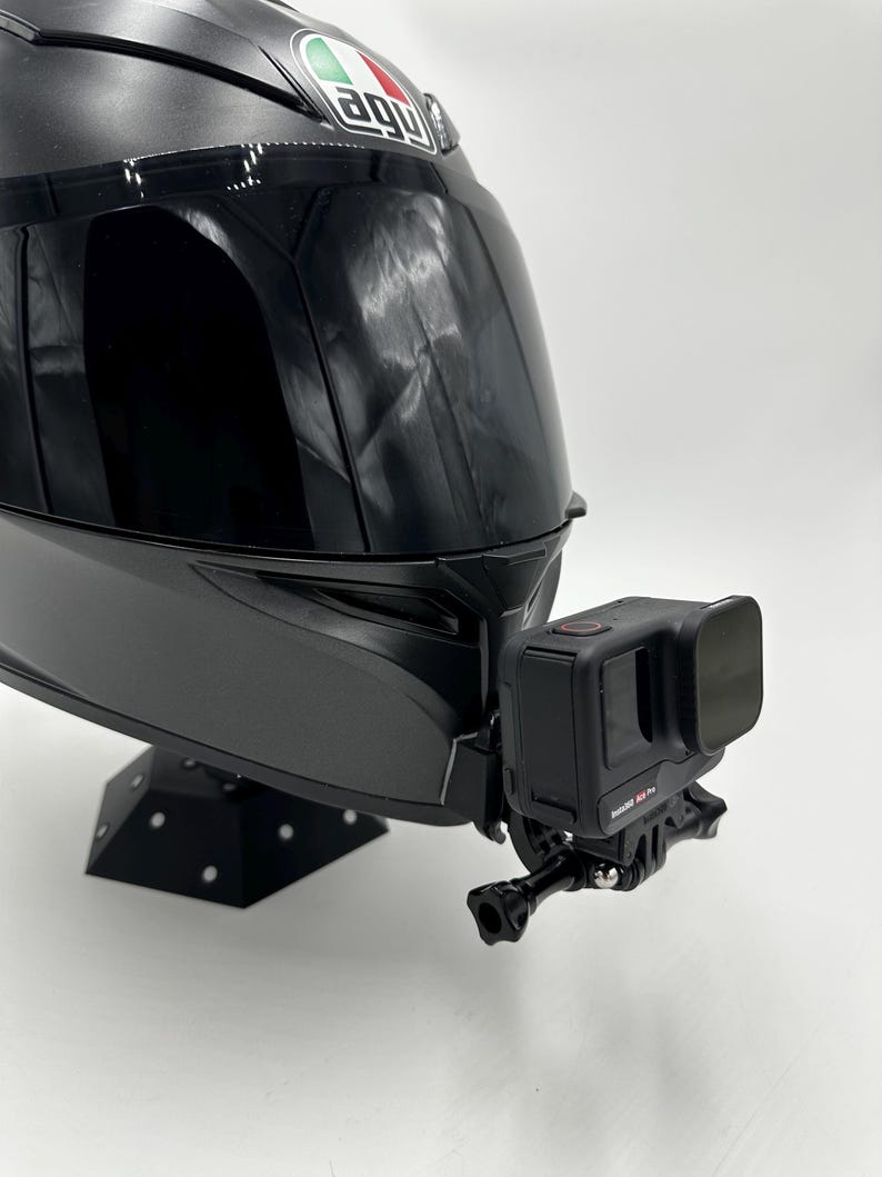May include: A black motorcycle helmet with a mounted black action camera. The helmet has a glossy finish and the camera is attached to the front. The helmet is on a black stand.