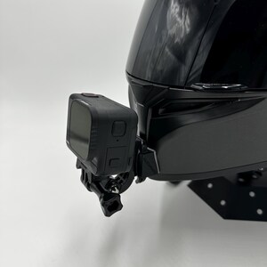 May include: A black motorcycle helmet with a black action camera attached to the side. The camera has a small screen and is mounted with a black bracket. The helmet is glossy and has a smooth surface.