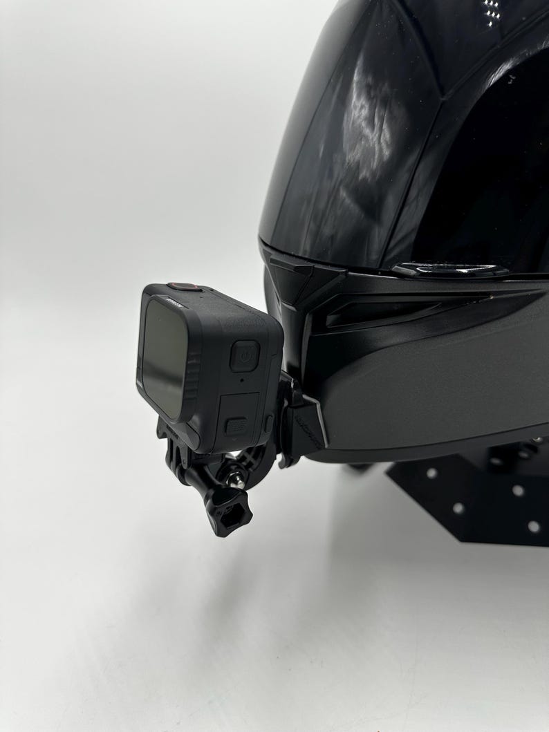 May include: A black motorcycle helmet with a black action camera attached to the side. The camera has a small screen and is mounted with a black bracket. The helmet is glossy and has a smooth surface.