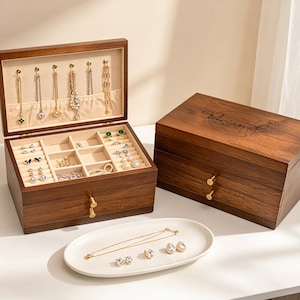 May include: Two wooden jewelry boxes, one open and one closed, sit on a white surface. The open box displays necklaces and rings. The closed box has the name "Vivian" and the date "10.10.2025" engraved on the lid. A white oval tray holds jewelry.