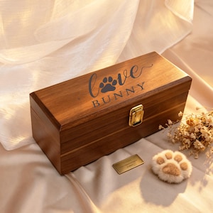 Könnte beinhalten: Eine rechteckige Holzkiste mit der Aufschrift "Love Bunny" und einem Pfotenabdruck. Sie hat einen goldenen Verschluss und eine kleine goldene Plakette. Ein plüschiges pfotenförmiges Objekt liegt daneben.