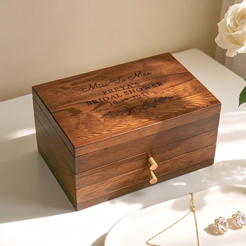 May include: A wooden jewelry box with a warm brown finish. The box is engraved with the text "Miss To Mrs FREYA'S BRIDAL SHOWER 10.19.2025" and floral accents. It has two small gold-colored knobs and a layered design.