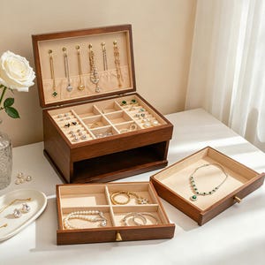 May include: A wooden jewelry box with multiple compartments and a hinged lid, showcasing various necklaces, rings, and earrings. The box is open, revealing a beige interior and a selection of gold and silver jewelry. Additional trays with jewelry are also visible.