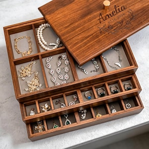 May include: A wooden jewellery box with multiple compartments, filled with various necklaces, bracelets, earrings, and rings. The lid is engraved with the name "Amelia" and the date 07.16.2026. The box is made of dark wood and has a gold-coloured knob.