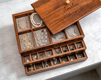 May include: A wooden jewelry box with multiple compartments, filled with various necklaces, bracelets, earrings, and rings. The lid is engraved with the name "Amelia" and the date 07.16.2026. The box is made of dark wood and has a gold-colored knob.