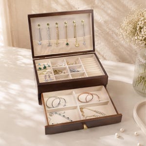 May include: A dark brown wooden jewelry box with multiple compartments and drawers. The top section displays necklaces and the lower drawer contains bracelets and rings. The interior is lined with a light beige fabric.