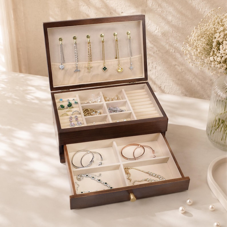 May include: A dark brown wooden jewelry box with multiple compartments and drawers. The top section displays necklaces and the lower drawer contains bracelets and rings. The interior is lined with a light beige fabric.