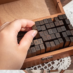 May include: A wooden box filled with individual letter and number stamps. A hand is holding up a stamp with the letter "G" on it. The stamps are dark brown and the box is a lighter brown color.