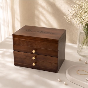 May include: A dark brown wooden jewelry box with three drawers and gold-colored knobs. The top of the box has the name "Lariah" engraved on it. The box is sitting on a white surface with a vase of flowers in the background.