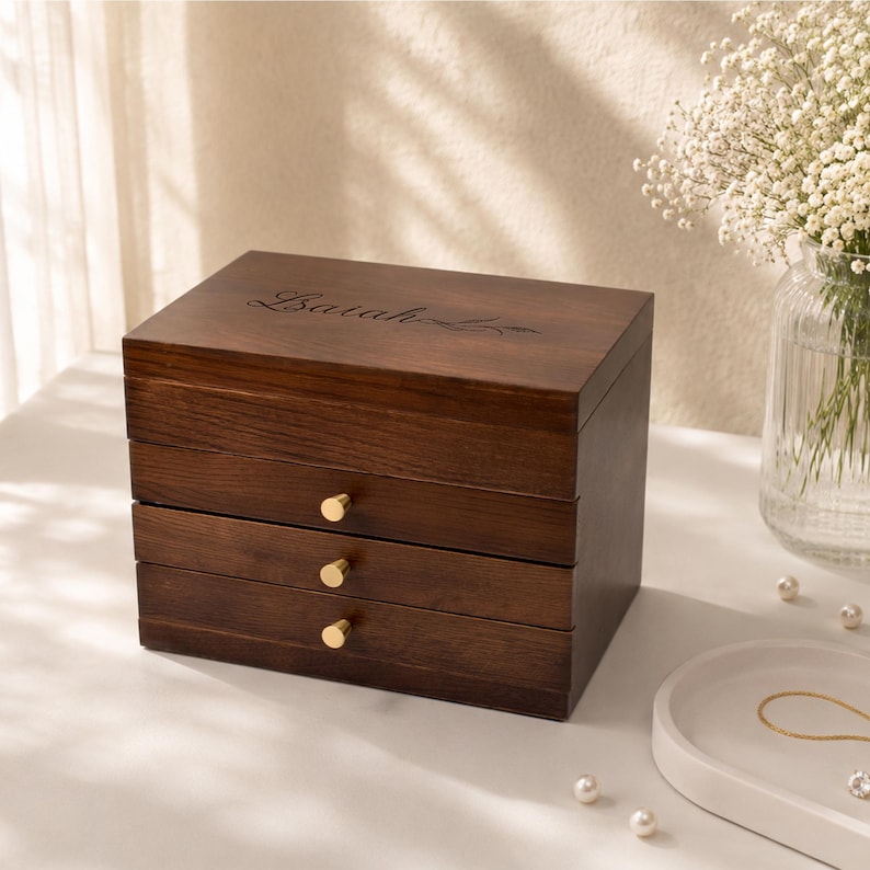 May include: A dark brown wooden jewelry box with three drawers and gold-colored knobs. The top of the box has the name "Lariah" engraved on it. The box is sitting on a white surface with a vase of flowers in the background.