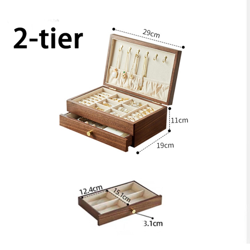 May include: A two-tier wooden jewelry box, 29cm wide, with a drawer and a hinged top. The interior is lined with a light-colored fabric and features compartments for rings, necklaces, and other jewelry. The drawer is 19cm deep and 11cm high.