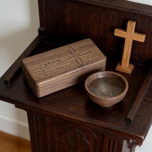 May include: A wooden box with a cross design and the name "Whitney" engraved on the lid, along with a small wooden cross and a wooden bowl. The items are displayed on a dark wooden stand.