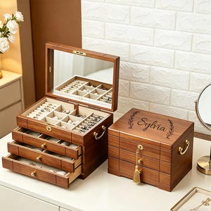 May include: Two wooden jewellery boxes with multiple drawers and compartments. One box is open, revealing jewellery, and has a built-in mirror. The other box is closed, with the name "Sylvia" engraved on the lid. Both boxes have gold-coloured handles.