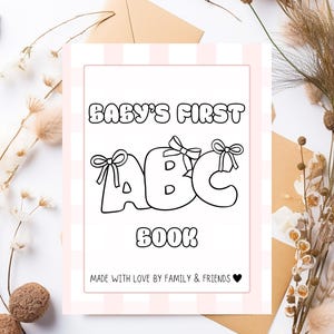 Op de afbeelding: Een baby's eerste ABC-boek met een roze en witte gestreepte rand. De titel staat in grote, vette letters, waarbij elke letter van het ABC is versierd met een strik. De onderkant van het boek luidt "Made with love by family & friends."