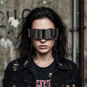 May include: A person wearing futuristic silver visor sunglasses. The sunglasses have a horizontal, rectangular design that covers the eyes. The person is wearing a black denim jacket and a black t-shirt. The background is a textured concrete wall.